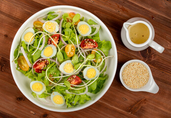 Big white bowl of lettuce, sprout salad with boil egg, onion and tomato mixed with Japanese sesame oil dressing on brown wooden table together with small olive oil jar and sesame cup