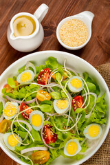 Big white bowl of lettuce, sprout salad with boil egg, onion and tomato mixed with Japanese sesame oil dressing on brown wooden table together with small olive oil jar and sesame cup