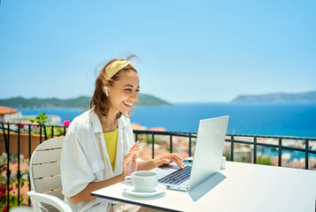 beautiful caucasian woman in wireless earphones talk speak on video call on computer while sitting at balcony with seaview on resort. female have online conversation, work chat conference or