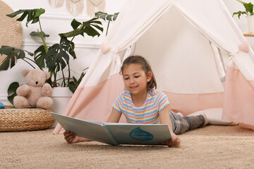 Cute little girl reading book near toy wigwam at home © New Africa