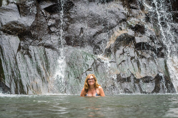 A pretty blonde woman swimming in a river under waterfall. Cute female traveler bathing in a beautiful lake under rocks and streams.