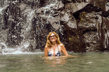 A pretty blonde woman sitting in a warm lake under waterfall and smiling happily. Travel and enjoy freedom after retirement. Female person on pension in tropical jungle.