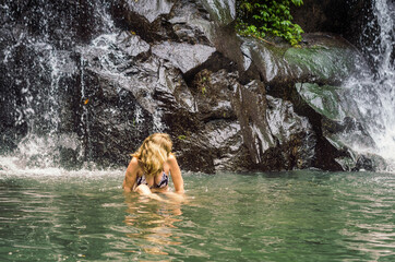 Anonymous blonde woman bathing in the river near tropical waterfall during summer hiking trip.