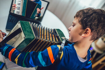 Focused boy playing accordion and watching online course on laptop while practicing at home. Online...