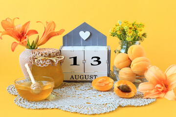 Calendar for August 13 : the name of the month of August in English, the number 13, flowers in vases, a jar of jam, apricots on a gray napkin, yellow background, side view