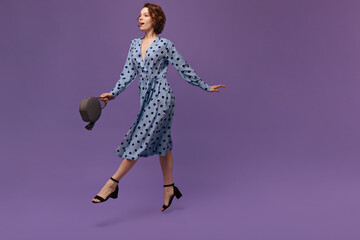 Young girl in dress jumping at studio. Pretty ginger-haired lady wearing polka dot outfit and black shoes, holding bag, looking aside and stretching legs against purple background