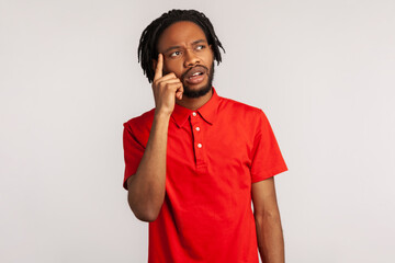 Portrait of young attractive bearded man with dreadlocks wearing red casual style T-shirt, thinking, has new idea, looking way with pensive expression. Indoor studio shot isolated on gray background.