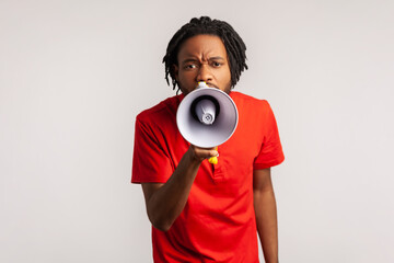 Man with dreadlocks wearing red casual style T-shirt, holding megaphone near mouth loudly speaking, screaming, making announcement, paying attention. Indoor studio shot isolated on gray background.
