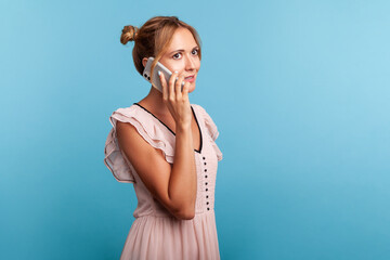 Mobile communication. Side view of good looking young adult woman in dress calling partner, talking on cell phone with calm expression. Indoor studio shot isolated on blue background.