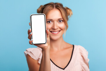 Portrait of blonde woman standing with charming smile, happy expression and showing mobile device, cell phone with mock up empty display to advertise. Indoor studio shot isolated on blue background.