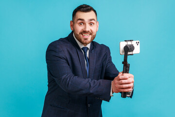 Naklejka premium Portrait of happy positive bearded businessman wearing official style suit streaming online, using smart phone and steadicam, broadcasting live stream. Indoor studio shot isolated on blue background.