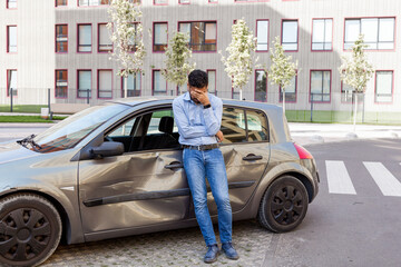 Fototapeta na wymiar Portrait of man wearing jeans and shirt standing in city street near broken car, covering face with palm, being despair after auto accident and smashed automobile. Outdoor shot.