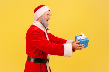 Side view of elderly man with gray beard wearing santa claus costume holding giving present box, greeting with Christmas and new year eve. Indoor studio shot isolated on yellow background.