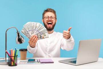 Happy excited bearded man showing thumbs up like gesture holding fan of dollar cash sitting at...