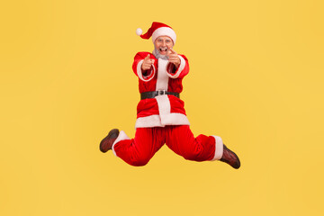 Elderly man with gray beard wearing santa claus costume jumping high and looking at camera, pointing with fingers, satisfied with winter holidays. Indoor studio shot isolated on yellow background.