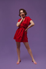 Full-length portrait of young girl isolated over violet background. Charming dark-haired lady with curly dark hairstyle, red dress and pink shoes, looking into camera and touching cheek