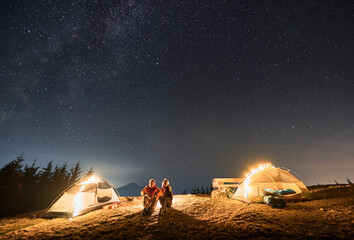 Young couple travelers sitting on grassy hill near tourist tents and looking at majestic night sky...