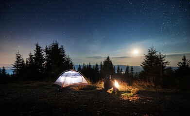 Side view of young man sitting on grass near his lighted tent and watching the night stars and moon shine. Recreation in mountains campsite outdoors. Beauty of lunar starry night sky.