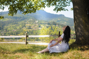 Young woman in white dress is sitting under a tree on mountain meadow.