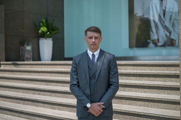 Portrait of handsome businessman in grey formal suit standing near modern building and looking at camera copy space. Serious face.