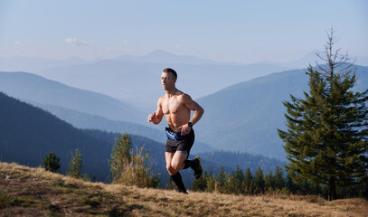 Naklejka na ściany i meble Professional athlete trail runner with well-developed muscles of torso running on mountain meadow in warm summer morning against backdrop of fabulous mountain beskids.
