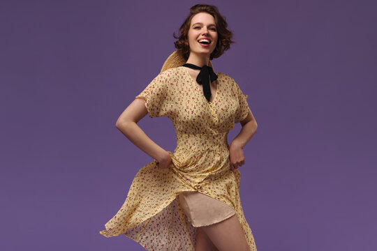 Photo in motion of young lady in floral yellow dress. Young ginger-haired girl wearing retro outfit, smiling and looking skirt against purple background