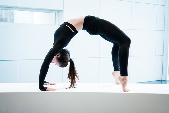 Young athletic flexible fitness woman practice chakrasana wheel, wearing black sportswear, in panoramic light window at home studio indoor on white background