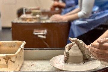 Hands of a ceramist while she is working on a potter's wheel against the background of other people working with clay