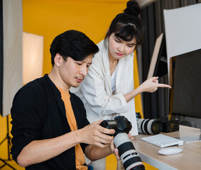 An unhappy male professional photographer working with young irritable female model by checking pictures together on the camera screen compared with photos on the computer monitor and found problems