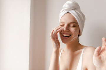 Joyful lady making washing and skincare procedures. Pale young girl with white towel on head and closed eyes, laughing while cleansing face, isolated over light background - Powered by Adobe