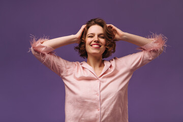 Naklejka premium Lounge and home wear concept. Portrait of charming young lady with dark curly hair, silk pajama, narrowed eyes, smiling and touching head isolated over violet background