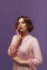 Lounge and wellness concept. Portrait of charming young ginger-haired lady, wearing pink pajama with feathers, looking into camera and posing isolated over purple background