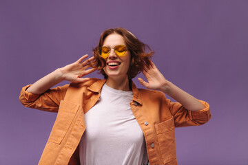 Expressive teenage girl dancing against violet background. Cute young dark-haired lady with white top and orange shirt, stylish bright sunglasses, smiling and moving