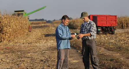 Farmer and buyer  holding Euro banknote with corn field with tractor and combine in background - obrazy, fototapety, plakaty