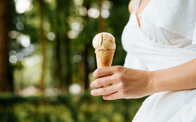 Close-up young woman in white summer dress holds vanilla ice cream in a waffle cone outdoors, selective focus on female hand. Hot weather, summer lifestyle concept.