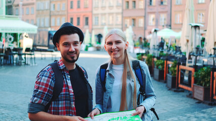 Happy young couple of tourists checking map and then smiling to the camera. They standing on a big market square of old european city.