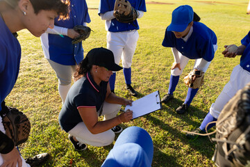 Diverse group of female baseball players in huddle around squatting female coach with clipboard