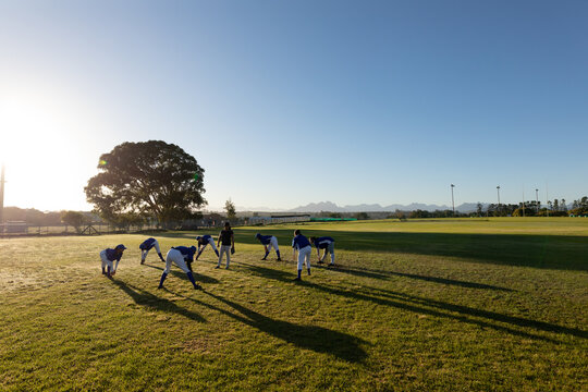 Diverse group of female baseball players with coach warming up in sunny field, stretching - Powered by Adobe