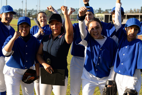 Diverse group of happy female baseball players and coach celebrating on baseball field after game