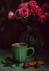 a bouquet of scarlet roses in a transparent vase on a gray table; next to it there is a green ceramic mug, in which a red rose bud is floating. Romantic setting, greeting card