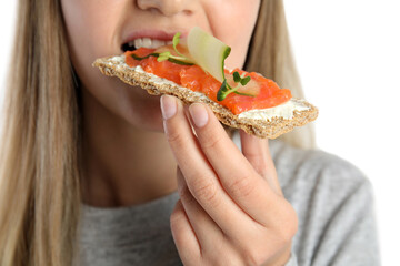 Woman eating fresh rye crispbread with cream cheese, salmon and cucumber on white background, closeup