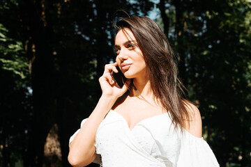 Portrait of a beautiful young Indian woman in a white summer dress talking on a mobile phone outdoors on a sunny day. Attractive smiling lady listening to someone on the phone