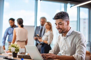 Young businessman working on his laptop in office. Young african executive sitting at his desk surfing internet on laptop computer. Confident young man at his desk