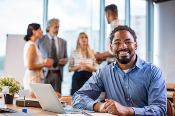Businessman Using Laptop At Desk In Busy Office.  Black businessman with colleagues sitting in a...