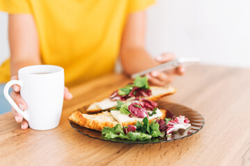 Young blonde woman in yellow t-shirt having breakfast with healthy sandwiches croissants and tea morning coffee in mug and using mobile phone in bright kitchen at home