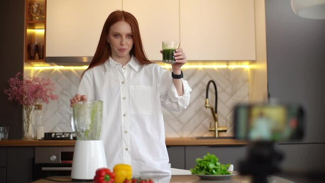 Attractive female blogger influencer using camera streaming and recording vlog video about benefit of drinking vegetarian juice. Happy young woman doing fresh smoothie during recording video.