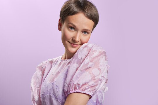 Shy beautiful caucasian young lady with light short hair in voluminous pastel pink dress. Emotion concept. Watching at the camera isolated over pastel purple background studio portrait medium shot