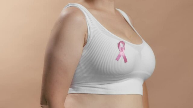 Busty girl in a seamless white bra rotate to show pink ribbon sign on her chest to raise awareness and support women fighting breast cancer. Anonymous slow motion studio shot video on beige background
