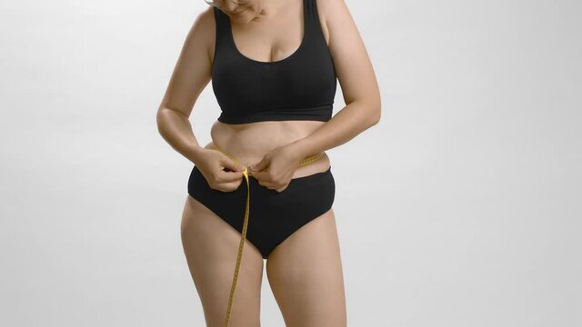 Plus size young caucasian woman in black underwear wants to know her waist volume using measure tape. White background still shot high quality studio video. Anonymous cowboy shot.