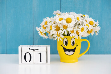 August 1st date on calendar and bouquet of chamomile in yellow mug with funny face against blue...